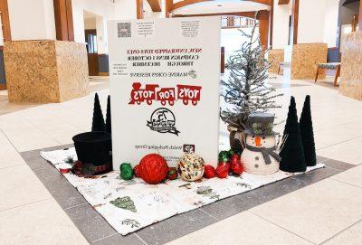 A tall white box with a red train logo that says Toys for Tots, surrounded by Christmas ornaments, small trees, and a snowman decoration.